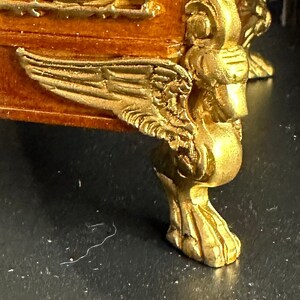 May include: A gold-painted wooden furniture leg with intricate carvings and a winged design. The leg is on a dark surface.
