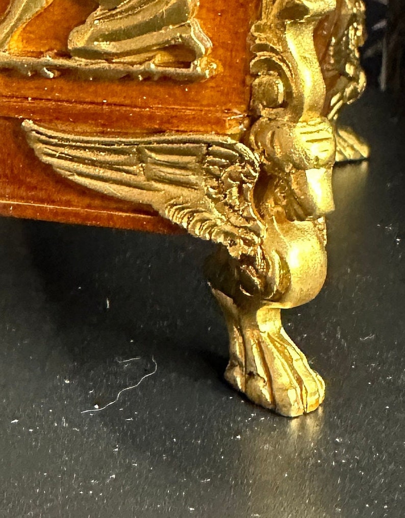 May include: A gold-painted wooden furniture leg with intricate carvings and a winged design. The leg is on a dark surface.