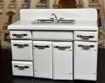 May include: White miniature kitchen sink and cabinet set with silver handles. The sink has two basins and a chrome faucet. The cabinet has three drawers and three doors.