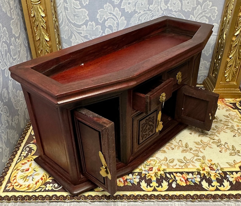 May include: A dark wood miniature cabinet with three drawers and two doors. The cabinet has a carved design on the front and brass handles. The cabinet is open and the drawers and doors are partially open.