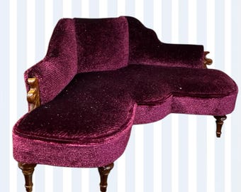 May include: Miniature burgundy velvet chaise longue with wooden legs and arm accents. The small sofa has a curved back and a rounded chaise end. A decorative piece for a dollhouse or miniature scene.