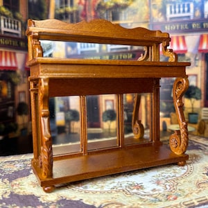 Peut inclure: Miniature vitrine en bois avec dos miroir et panneaux de verre. Le meuble présente des détails sculptés et une finition marron foncé. La conception comprend une étagère et des volutes décoratives, parfait pour la décoration de maison de poupée.