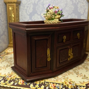May include: A dark wood cabinet with two doors and three drawers. The cabinet has gold-colored hardware and a decorative carved design on the front. A small basket of flowers sits on top of the cabinet.