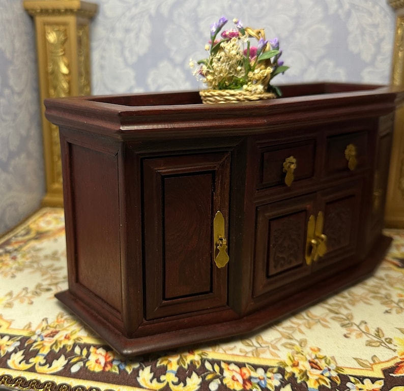 May include: A dark wood cabinet with two doors and three drawers. The cabinet has gold-colored hardware and a decorative carved design on the front. A small basket of flowers sits on top of the cabinet.