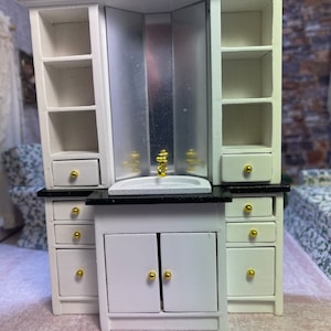 May include: White miniature bathroom vanity with a black countertop, a small sink, and gold hardware. The vanity has two doors and three drawers on each side. The vanity is part of a larger set that includes a mirror and shelves.