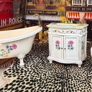Puede incluir: Un juego de baño que incluye una bañera con patas de garra, un tocador y un inodoro. Los elementos de porcelana blanca están decorados con diseños florales rosas y verdes. El tocador tiene una encimera de mármol y puertas de armario. El juego está sobre una alfombra con estampado de leopardo.