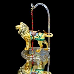 May include: A carousel lion figurine with a tan body, gold mane, and a brown saddle. The lion is mounted on a clear, circular platform with a red and gold pole. The background is black.