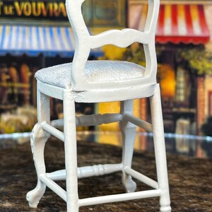 May include: White wooden bar stool with a padded seat and a decorative back. The stool has a simple design with a rectangular base and four legs.