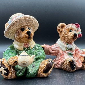 Puede incluir: Dos osos de peluche de cerámica que llevan vestidos y sombreros, uno sosteniendo una tetera y el otro una taza de té. Los osos están sentados sobre una superficie oscura.