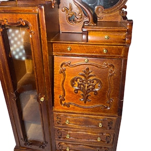 May include: A miniature wooden vanity with a mirror, drawers, and a cabinet. The vanity is a rich brown color with ornate carvings and gold-colored hardware. The cabinet door is open, revealing a glass-fronted display area.