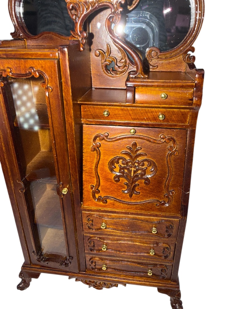 May include: A miniature wooden vanity with a mirror, drawers, and a cabinet. The vanity is a rich brown color with ornate carvings and gold-colored hardware. The cabinet door is open, revealing a glass-fronted display area.