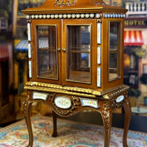 May include: A miniature wooden display cabinet with glass doors and ornate details. The cabinet has a gold trim and painted floral accents. It sits on four carved legs and is displayed on a patterned rug.