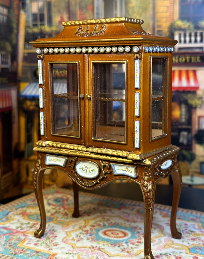 May include: A miniature wooden display cabinet with glass doors and ornate details. The cabinet has a gold trim and painted floral accents. It sits on four carved legs and is displayed on a patterned rug.