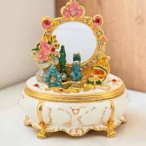 Puede incluir: Una caja de joyería decorativa y ornamentada con un espejo de tocador. La caja blanca tiene adornos y patas doradas, y presenta un pequeño espejo enmarcado en oro y rosa. La parte superior de la caja tiene figuras en miniatura y un pequeño ramo de flores rosas.