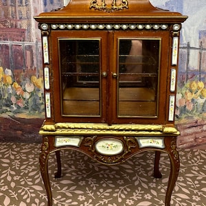 May include: A brown wooden display cabinet with glass doors and ornate gold trim. The cabinet has a decorative floral design painted on the sides and a floral design on the front. The cabinet sits on four ornate legs.