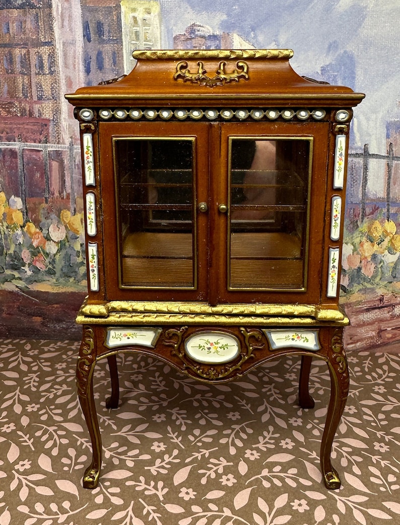 May include: A brown wooden display cabinet with glass doors and ornate gold trim. The cabinet has a decorative floral design painted on the sides and a floral design on the front. The cabinet sits on four ornate legs.