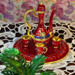 Puede incluir: Un juego de té de metal rojo y dorado con una tetera, tres tazas y una bandeja. La tetera tiene un diseño decorativo y joyas azules. Las tazas tienen bordes dorados y joyas azules.
