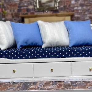 Op de afbeelding: Miniatuur wit houten bank met drie lades en gouden knoppen. Het heeft een marineblauw stippelkussen en drie kussens: twee blauwe en een wit gestreept. De bank maakt deel uit van een poppenhuismeubelset.