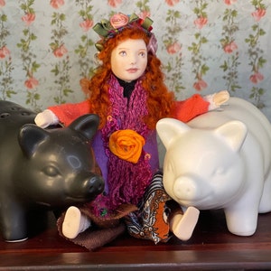 May include: A doll with red hair wearing a floral crown and a purple and orange patterned dress sits with two piggy banks, one black and one white.