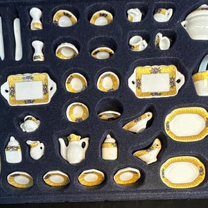 Op de afbeelding: Een miniatuur porseleinen theeservies in een zwarte lade met gouden rand. De set bevat een theepot, roomkan, suikerpot, kopjes, schoteltjes, borden en een serveerschaal. Het porselein is versierd met bloemmotieven en gouden accenten.
