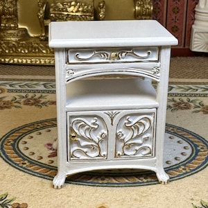 May include: A miniature white nightstand with gold accents. The nightstand features a drawer, an open shelf, and a cabinet with ornate floral designs. The cabinet doors have gold knobs. The nightstand is a decorative piece.