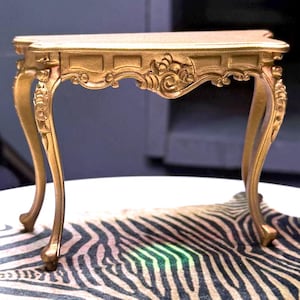 May include: A small, ornate gold-colored table with curved legs and decorative carvings. The table has a rectangular top and is set on a zebra-print surface. The table is approximately 12 inches long.