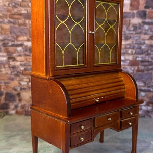 May include: A wooden roll top desk with a glass-front cabinet above. The desk has a roll top lid, four drawers, and four legs. The cabinet has two glass doors with a decorative gold trim.