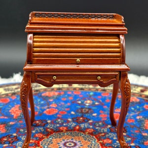 May include: A miniature wooden roll top desk with a brown finish and ornate legs. The desk is sitting on a blue and red patterned rug.