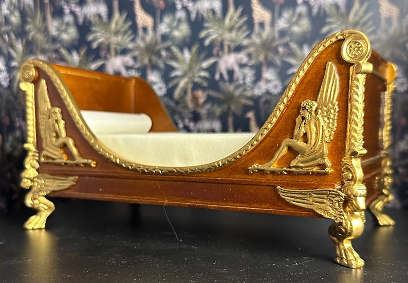 May include: A wooden chaise lounge with gold accents and winged cherub figures. The chaise has a white fabric cushion and sits on four gold feet.
