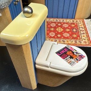 Puede incluir: Un inodoro blanco en miniatura con una tapa amarilla y una revista en el asiento. El inodoro está unido a una pared de madera con tablones pintados de azul y blanco. Una alfombra de patrón rojo y crema está en el suelo.
