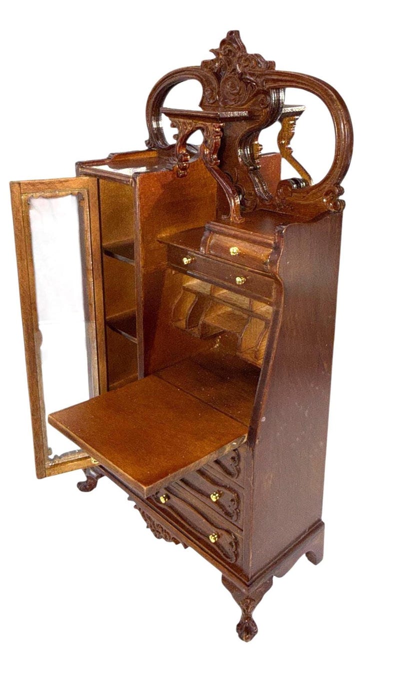 May include: Miniature wooden secretary desk with a dark brown finish. The desk features a drop-down writing surface, drawers with gold-colored knobs, and a glass-doored cabinet. The top has an ornate, arched design.