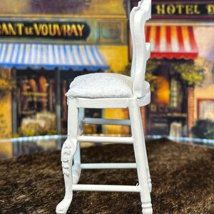 May include: White wooden chair with a white cushioned seat and a carved design on the legs. The chair is on a brown surface.