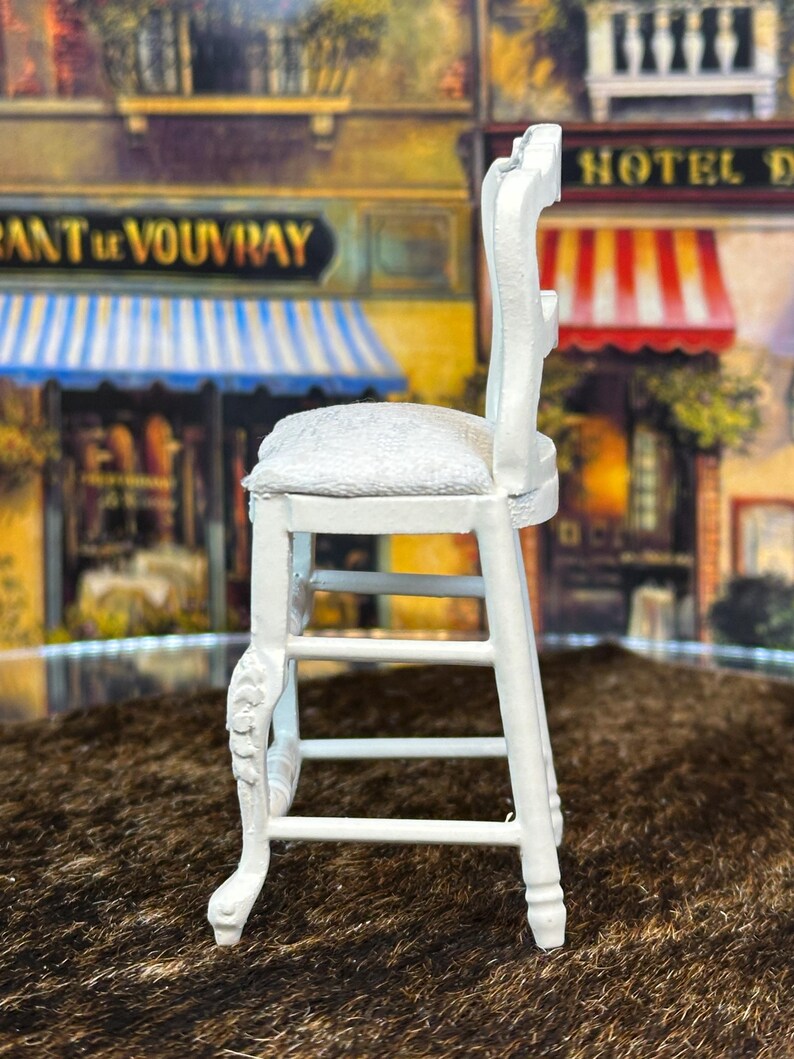 May include: White wooden chair with a white cushioned seat and a carved design on the legs. The chair is on a brown surface.