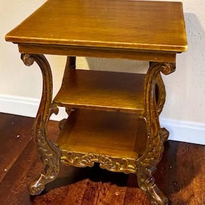 May include: A brown wooden side table featuring a square top, a middle shelf, and a lower shelf. The table is supported by four curved legs with intricate carved details and scrollwork, finished with a smooth, polished wood grain surface.