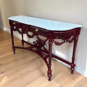 May include: A dark red console table featuring a light blue speckled top, two drawers with small gold knobs, and intricate carved detailing along the apron and legs. The piece stands on four slender legs connected by a central stretcher.