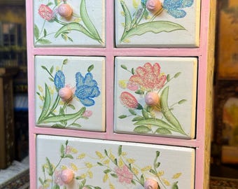 Könnte beinhalten: Eine rosa Holzkistenkommode mit fünf Schubladen. Jede Schublade ist mit einem handgemalten Blumendesign verziert, das rosa, blaue und gelbe Blumen zeigt. Die Schubladen haben kleine Holzknöpfe. Die Kommode steht auf einem gemusterten Teppich. Der Text "HOME & HEART COLLECTION" ist auf einem herzförmigen Etikett sichtbar.