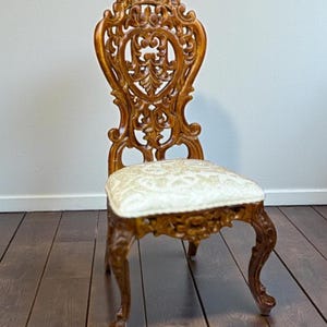 May include: A wooden chair with a high, intricately carved backrest featuring scrollwork and floral motifs. The seat is upholstered in a cream-colored fabric with a subtle, raised damask pattern. The legs are curved and feature carved details.