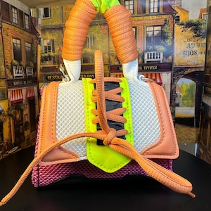 May include: A small pink, white, and neon green handbag with a mesh design and orange handles. The bag has a black and orange shoelace that is tied into a bow.