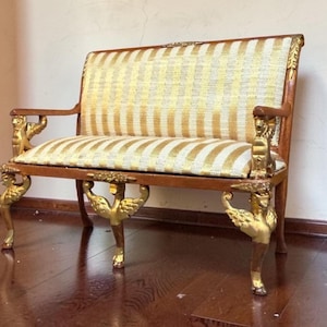 May include: An antique loveseat with a wooden frame and gold accents. The seat and back are upholstered in a striped pattern of gold and cream fabric. The legs and arm supports feature ornate gold detailing.