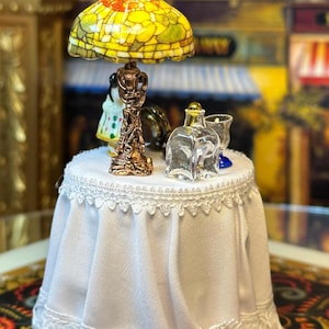 May include: Miniature table with a white lace tablecloth, holding a stained glass lamp, a small figurine, and a clear glass bottle. The lamp has a yellow and red shade. A miniature scene.