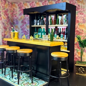 Puede incluir: Un conjunto de bar en miniatura con una barra negra, cuatro taburetes de bar negros y un armario de bar negro con estantes de vidrio llenos de botellas de licor en miniatura. El bar está instalado en un entorno de casa de muñecas con un fondo de papel tapiz floral.