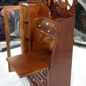 May include: Miniature wooden desk with a dark brown finish. The desk features a drop-down writing surface, drawers with gold-colored knobs, and an ornate mirror. The desk has a cabinet with a glass door and decorative carvings.