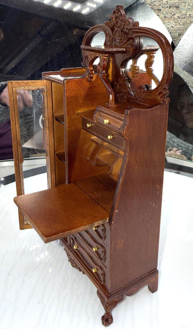 May include: Miniature wooden desk with a dark brown finish. The desk features a drop-down writing surface, drawers with gold-colored knobs, and an ornate mirror. The desk has a cabinet with a glass door and decorative carvings.