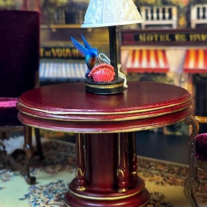 May include: A miniature red wooden table with a gold trim and a white lamp with a floral design shade. The lamp is topped with a blue hummingbird figurine and a pink and white shell.