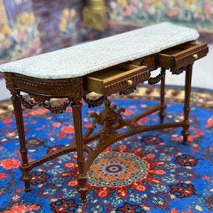 May include: Miniature brown wooden console table with two drawers open. The table has a white and gray marble top and ornate carved details. The drawers have gold knobs. The table is on a blue rug with a floral pattern.