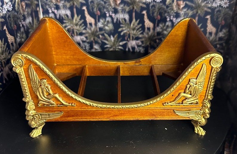 May include: A wooden decorative shelf with a gold finish. The shelf is shaped like a chaise lounge with winged cherubs on each side. The shelf has a gold rope design around the edge.