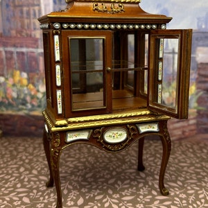 May include: A miniature wooden display cabinet with glass doors and shelves. The cabinet has a brown finish with gold accents and floral details. It is decorated with white beads and has a decorative top.