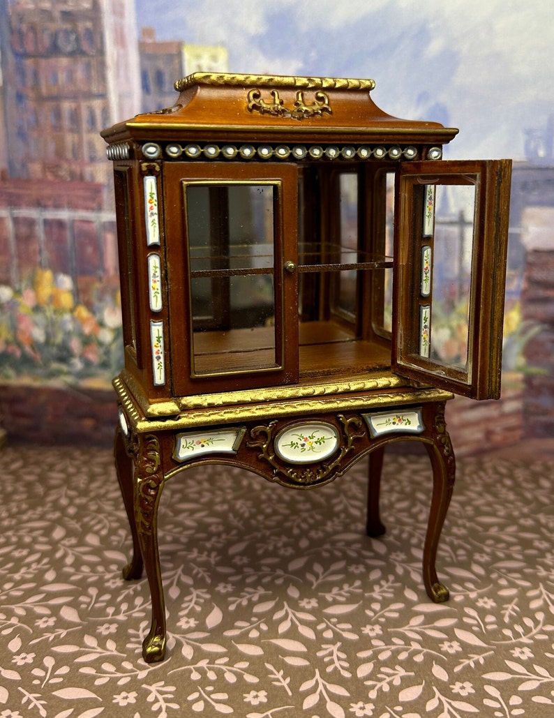 May include: A miniature wooden display cabinet with glass doors and shelves. The cabinet has a brown finish with gold accents and floral details. It is decorated with white beads and has a decorative top.