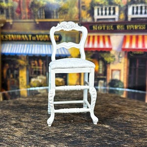 May include: White wooden chair with a white upholstered seat and a carved back. The chair has a simple design with four legs and a single rung.