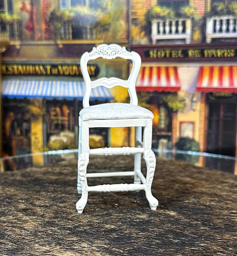 May include: White wooden chair with a white upholstered seat and a carved back. The chair has a simple design with four legs and a single rung.